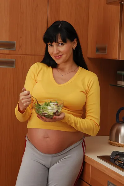 Jonge zwangere vrouw verse salade eten in een keuken. — Stockfoto