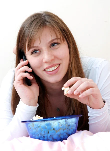 Hermosa joven disfrutando comiendo palomitas de maíz y hablando por teléfono celular en su cama . — Foto de Stock