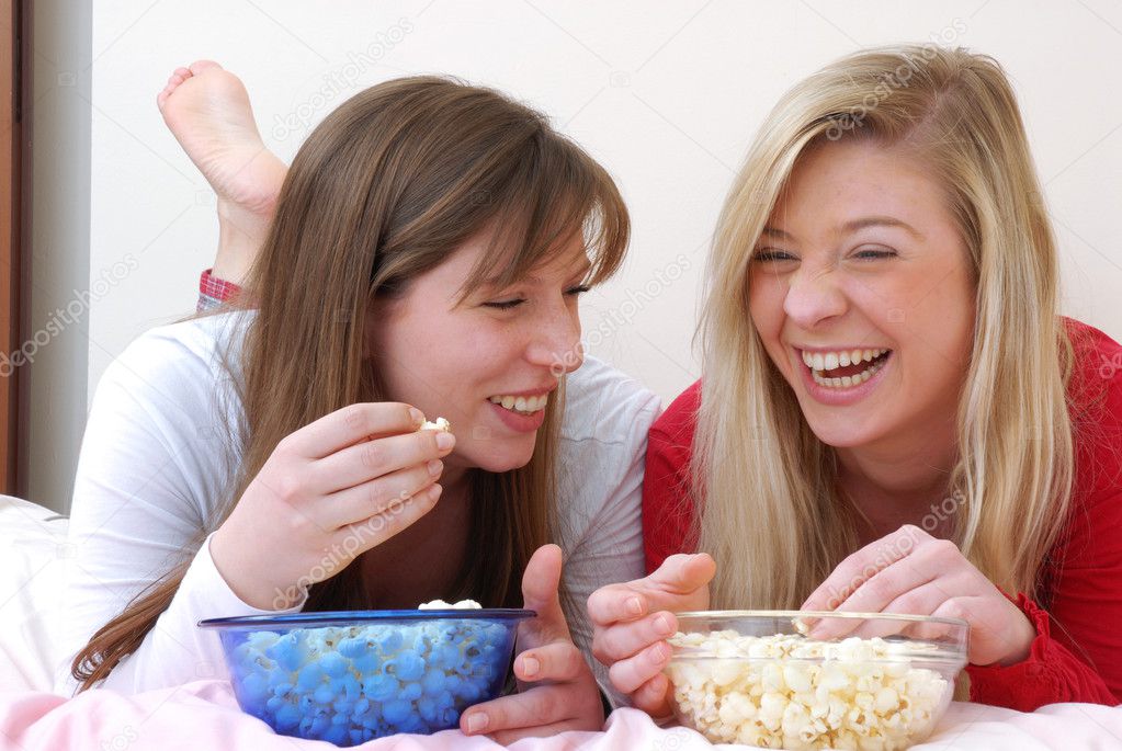 Two young women eating popcorn on bed.