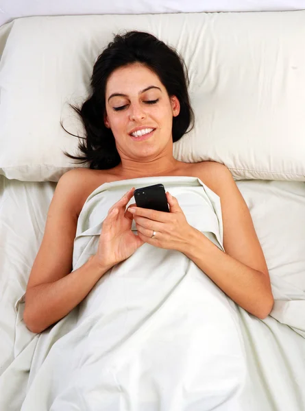 若い女性がベッドの中で携帯電話上でチャット — ストック写真