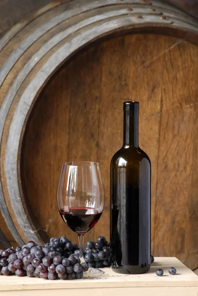 Бутылка красного вина, чашка и виноград на фоне бочек . — стоковое фото
