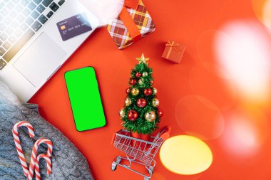 Kredi kartıyla bilgisayardan Noel alışverişi. Yeni yıl dekorasyonları, baston şekerler, alışveriş arabası ve hediye kutusunun üstü kırmızı arka planda yatıyordu. Mutlu Noeller kış tatili satış konsepti