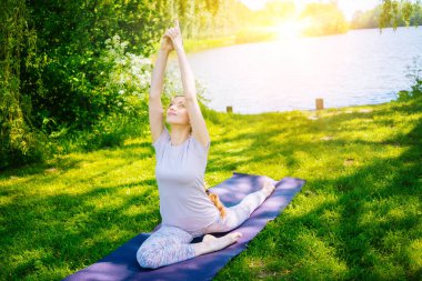 Parkta yoga yapan genç bir kadın. Kız yoga pozisyonunda egzersiz yapıyor. Nilüfer pozisyonunda oturup yoga yapan mutlu ve sağlıklı bir kadın. Gün batımında meditasyon ve spor.
