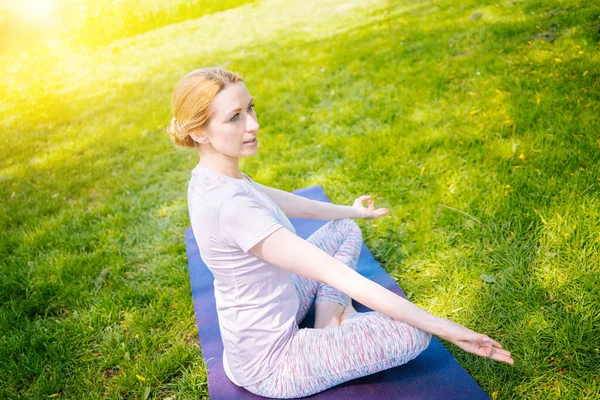 Mujer joven haciendo yoga asana en parque. chica estirando ejercicio en posición de yoga. mujer feliz y saludable sentada en posición de loto y practicando yoga. meditación y deporte al atardecer al aire libre. Imagen De Stock