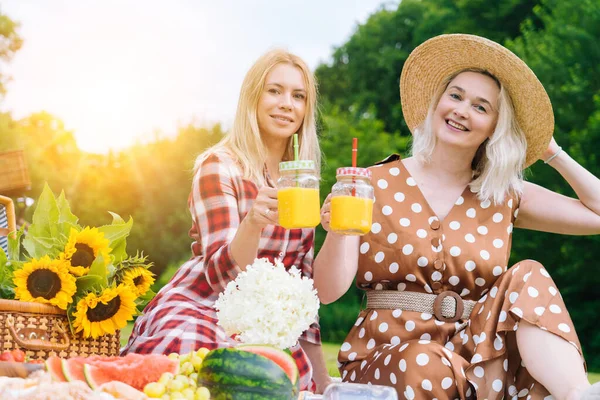 Friends is making picnic outdoor. Laughing girls sitting on white knit picnic blanket drinking wine. Happy young women talking, smiling outdoors on Summer picnic on sunny day at park