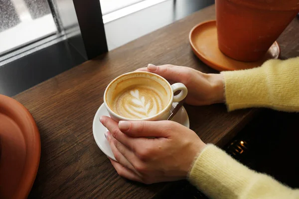 Manos de mujer en suéter amarillo sosteniendo taza de café fresco con arte latte —  Fotos de Stock