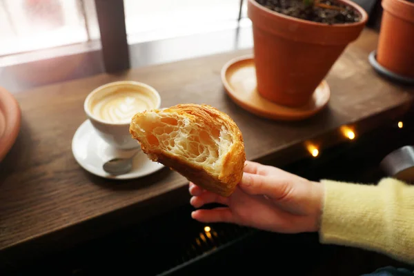 Main féminine tenant un croissant fraîchement mordu. Coupe de cappuccino sur fond — Photo