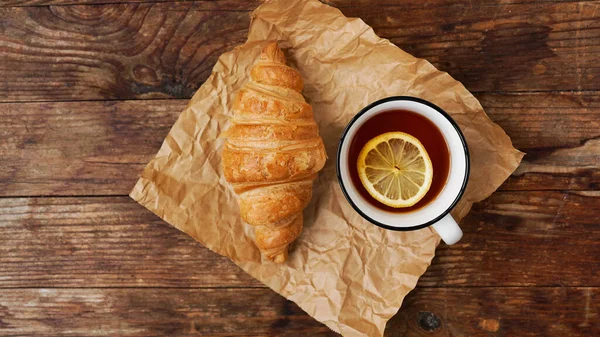 Fresh croissants and a cup of tea with lemon on a wooden table. Top view — Stock Photo, Image