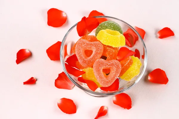 Gekleurde snoepjes met twee rode harten in glazen kom en rozenblaadjes — Stockfoto