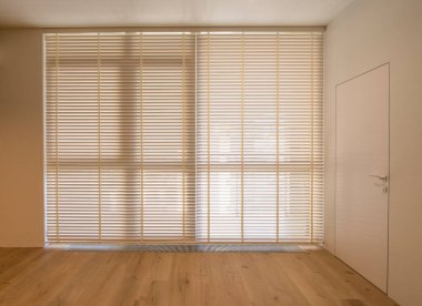 Motorized wood blinds in the interior. Automatic venetian blinds beige color on large windows. Wooden slats 50mm wide. A door to the room is near the window. 