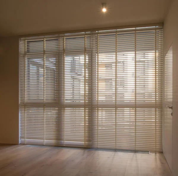 Motorized wood blinds in the interior. Automatic venetian blinds beige color on large windows. Wooden slats 50mm wide. A door to the room is near the window. 