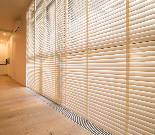 Motorized wood blinds in the interior. Automatic venetian blinds beige color on large windows. Wooden slats 50mm wide. Wood floor, beige walls.