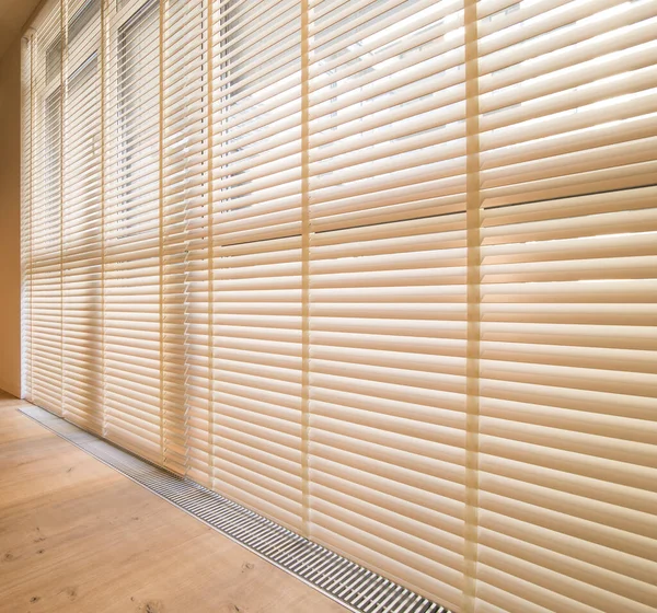 Motorized wood blinds in the interior. Automatic venetian blinds beige color on large windows. Wooden slats 50mm wide. Wood floor, beige walls.