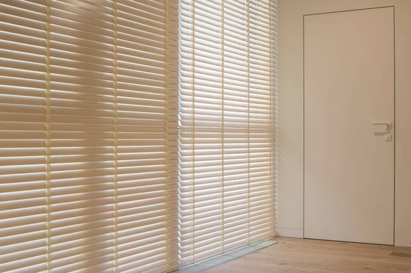 Motorized wood blinds in the interior. Automatic venetian blinds beige color on large windows. Coulisse wooden slats 50mm wide. A door to the room is near the window.