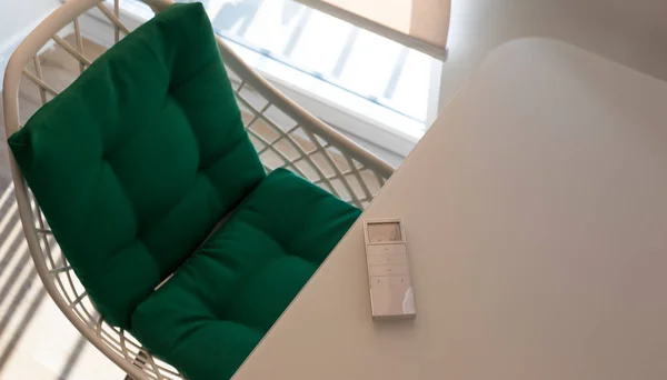 White remote control panel for automatic roller blinds lies on a beige table in the living room. Nearby is a chair with a green pillow. On the window, a roller blind with automatic control.