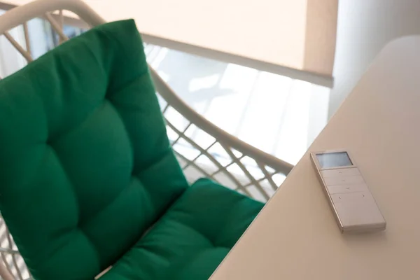 White remote control panel for automatic roller blinds lies on a beige table in the living room. Nearby is a chair with a green pillow. On the window, a roller blind with automatic control.