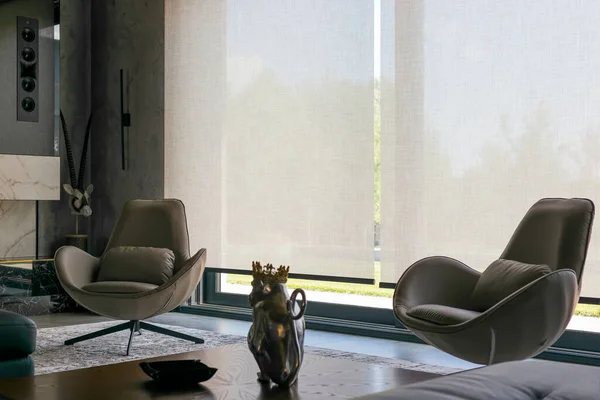 Interior design with roller blinds in the background. Automatic solar shades of large sizes on the window. Fabric with linen texture. In front of a large window is a chair on a carpet. 