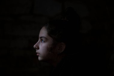 War in Ukraine. A child in a basement, looking at the camera. A beam of light falls on her face. Portrait of the little girl in the dungeon with dark background. Pray for Ukraine.