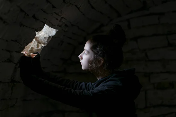 War in Ukraine. A child in a basement looking in a small window. A beam of light falls on her face. Portrait of the little girl in the dungeon with dark background. Pray for Ukraine. 