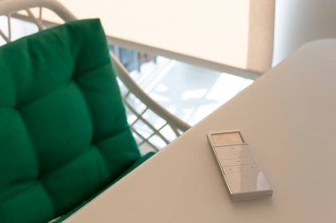 Otomatik patenler için beyaz uzaktan kumanda paneli oturma odasındaki bej bir masada yatıyor. Yakınlarda yeşil yastıklı bir sandalye var. Pencerede otomatik kontrollü bir paten var.. 