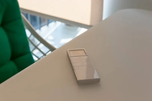 White remote control panel for automatic roller shades lies on a beige table in the living room. Nearby is a chair with a green pillow. On the window, a roller blind with automatic control.
