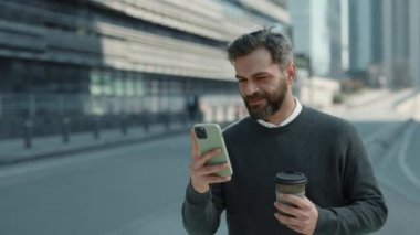 Gülümseyen işadamı elinde kahve ve cep telefonuyla yürüyor.