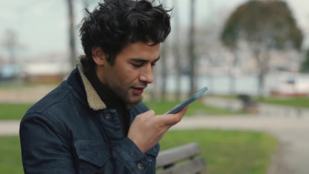 Mężczyzna siedzi w parku i używa telefonu komórkowego do przesyłania wiadomości dźwiękowych — Wideo stockowe