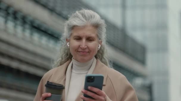 Idős hölgy fülhallgatóban, aki az utcán sétál és mobilt használ — Stock videók