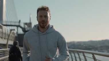 Gri kapüşonlu genç sakallı adam şehir sokaklarında koşuyor.