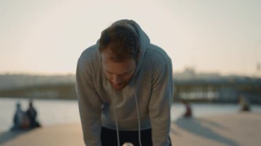 Spor kıyafetleri içinde yorgun bir adam akşam koşusundan sonra zor nefes alıyor ve şehir nehrinin yakınındaki kameraya bakıyor. Çalışmadan sonra enerji tazelemek için şehir nehrinin yanında duran genç sporcunun portresi..