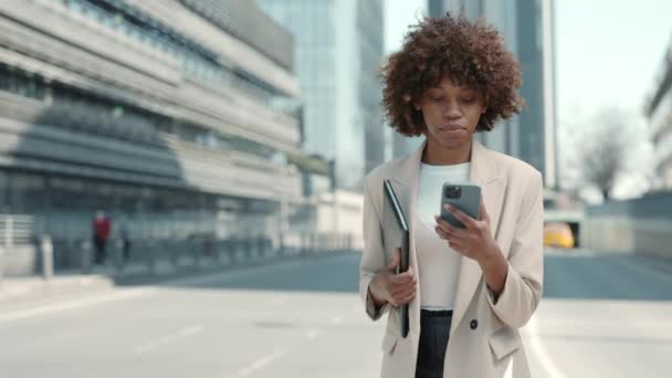 Mujer afroamericana confiada llevando documentos y usando el móvil al aire libre — Vídeo de stock