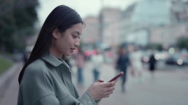 Şehir merkezinde elinde modern cep telefonuyla günlük giysiler içinde duran Asyalı güzel bir kadının portresi. Şehir caddesinde internette sörf yapan ve kameraya bakan mutlu bir kadın..