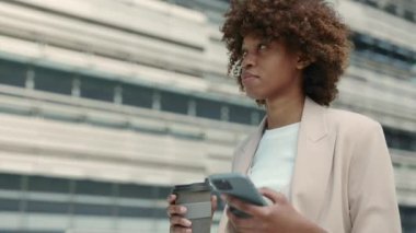 Kendine güvenen Afro-Amerikan kadın şık resmi kıyafetler giymiş akıllı telefon kullanıyor, kahve taşıyor ve şehir caddesinde yürüyor. Büyük şehirde iş kadını..