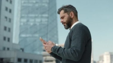 Sakallı, olgun bir adam. Şehir caddesinde akıllı telefon kullanıyor.