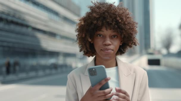 Retrato de mujer afroamericana en traje formal usando un teléfono celular personal mientras está de pie en la calle de la ciudad mirando la cámara. Mujer atractiva con el pelo rizado navegar por Internet en el teléfono inteligente al aire libre. — Vídeos de Stock