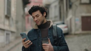 Cep telefonu, sırt çantası ve kahve taşıyan mutlu bir adam sokakta yürüyor.