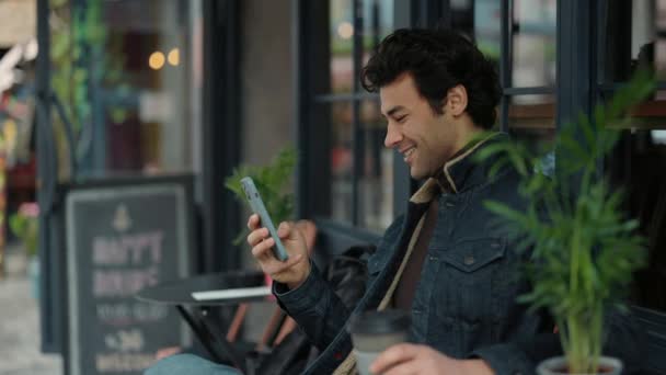 Chico alegre usando teléfono inteligente mientras descansa en la terraza de la cafetería — Vídeo de stock