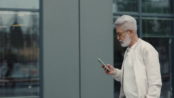 Starší muž pomocí mobilu při chůzi v blízkosti kancelářské budovy — Stock video