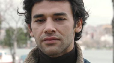 Kendine güvenen, siyah gözlü, dalgalı saçlı, şehir parkındaki kameraya bakan Arap bir adama yaklaş. Bir kulağına piercing takmış yakışıklı bir adamın açık hava portresi. Bulanık arkaplan.