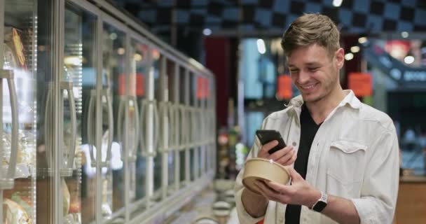 Mladý veselý muž držící produkt a kontrolující jeho cenu při použití smartphonu v supermarketu. Millennial pohledný muž usmívá při pohledu na obrazovce telefonu a dělá nákupy. — Stock video