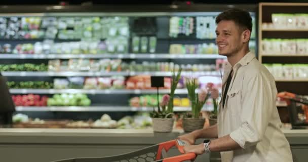 Wesoły młodzieniec uśmiechający się i pchający wózek podczas spaceru po supermarkecie. Milenium przystojny facet w dobrym nastroju robi zakupy. Zamazane półki spożywcze na tle.Koncepcja prawdziwego życia. — Wideo stockowe