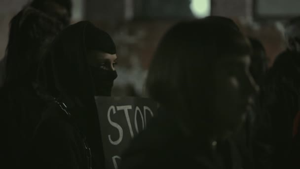 Lviv, Ukraine - June 3, 2020: Female person in black balaclava holding carton placard and raising up clintched fist with people crowd. Protestors at night city street. Concept of strike. — Stock Video