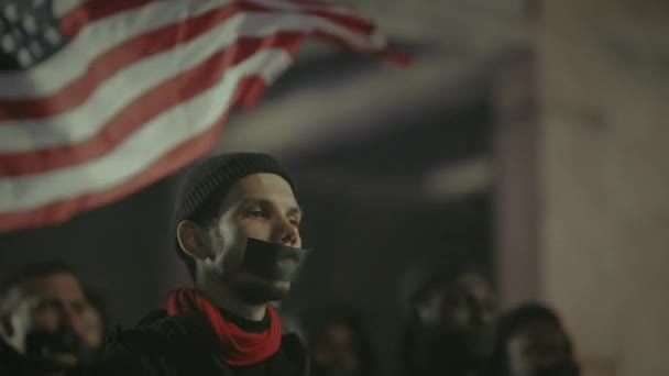 Pemuda Kaukasia dengan pita hitam di mulutnya berdiri di jalan kota malam. Kerumunan demonstran dengan bendera american di latar belakang. — Stok Video