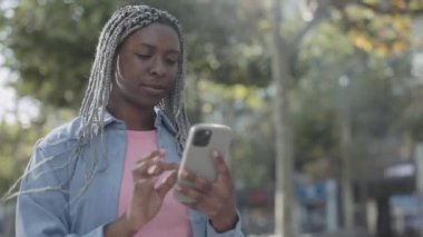 Afro örgülü bir kadın sokakta yürüyor ve cep telefonu kullanıyor.