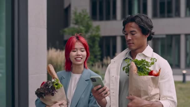 Pareja asiática usando móvil mientras camina con bolsas de comestibles — Vídeos de Stock