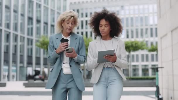 Wieloetniczne kobiety z telefonem komórkowym i tabletem spacerujące ulicą — Wideo stockowe