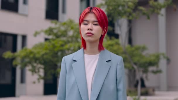 Young asian business woman standing on city street and looking at camera with serious facial expression. Female person having red hair and wearing blue jacket. — Stock Video