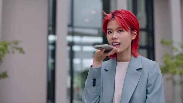 Pretty asian business woman using modern smartphone for recording audio message outdoors. Young red haired lady standing on city street and talking in microphone of cell phone. — Stock Video