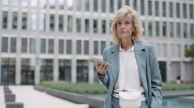 Şehir merkezinde elinde akıllı telefonuyla kameraya bakan olgun bir iş kadını portresi. Modern aletlerle internette sörf yapan kendine güvenen bir kadın..