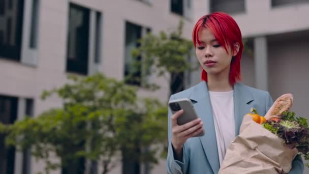 Mujer asiática usando móvil y sosteniendo bolsa de comestibles en la calle — Vídeo de stock